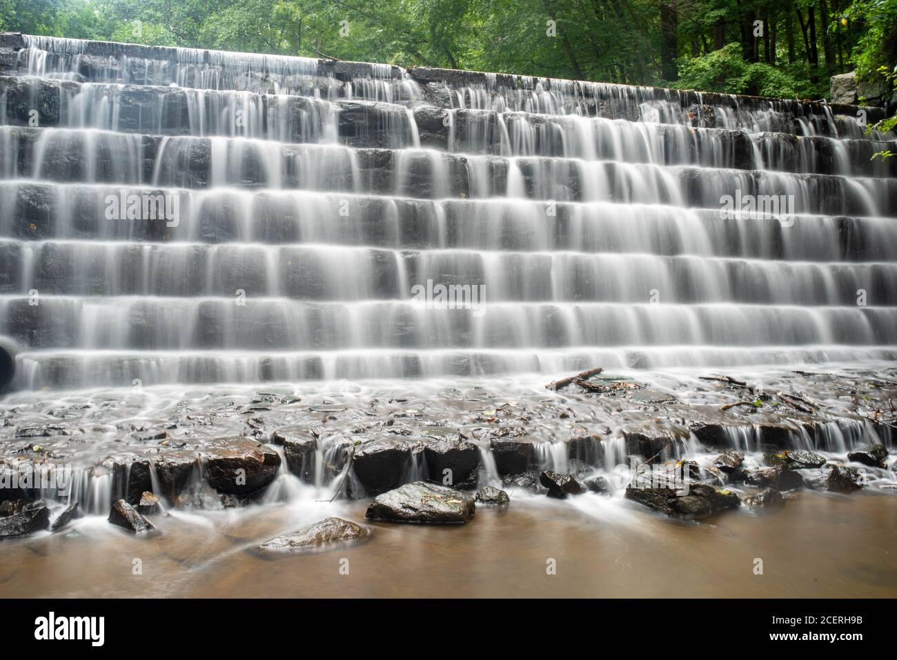 Immagine full frame a lunga esposizione di una cascata a più livelli in un canale d'acqua della foresta con fogliame verde dietro e acqua liscia in primo piano. Foto Stock