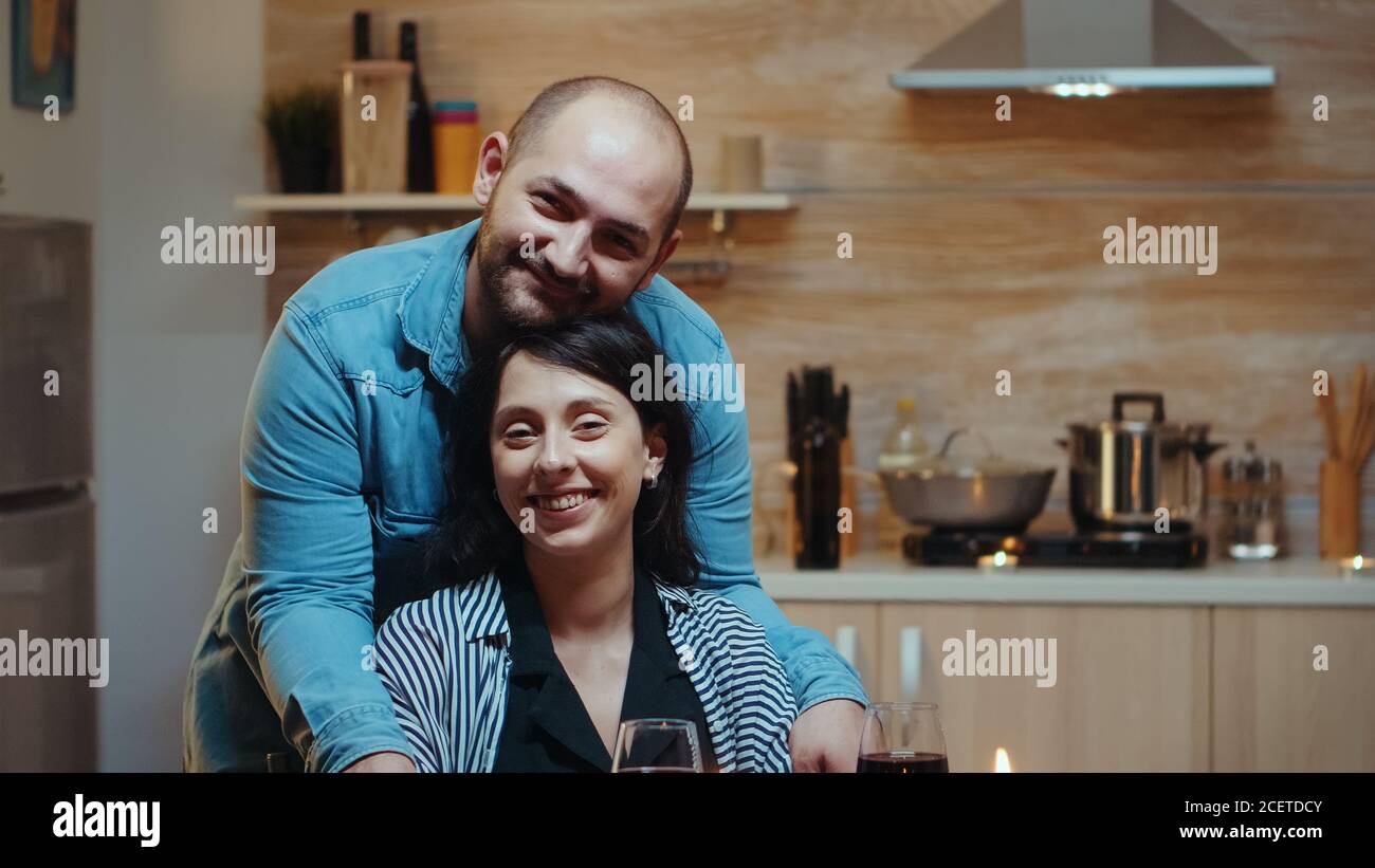 Portret di coppia allegra guardando la macchina fotografica, sorridente, seduto al tavolo in cucina durante la cena romantica. Felice poeple sposato momenti speciali teneri, injoryng il pasto a lume di candela celebrazione Foto Stock