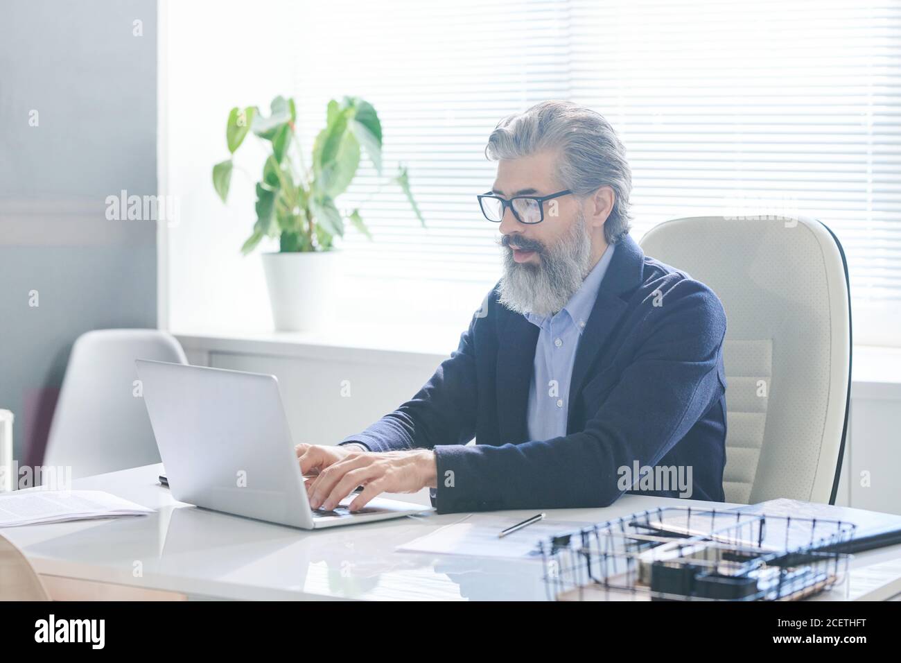 Ritratto di un operaio maschile concentrato che indossa occhiali seduti alla scrivania dell'ufficio che scriva qualcosa sul computer portatile Foto Stock