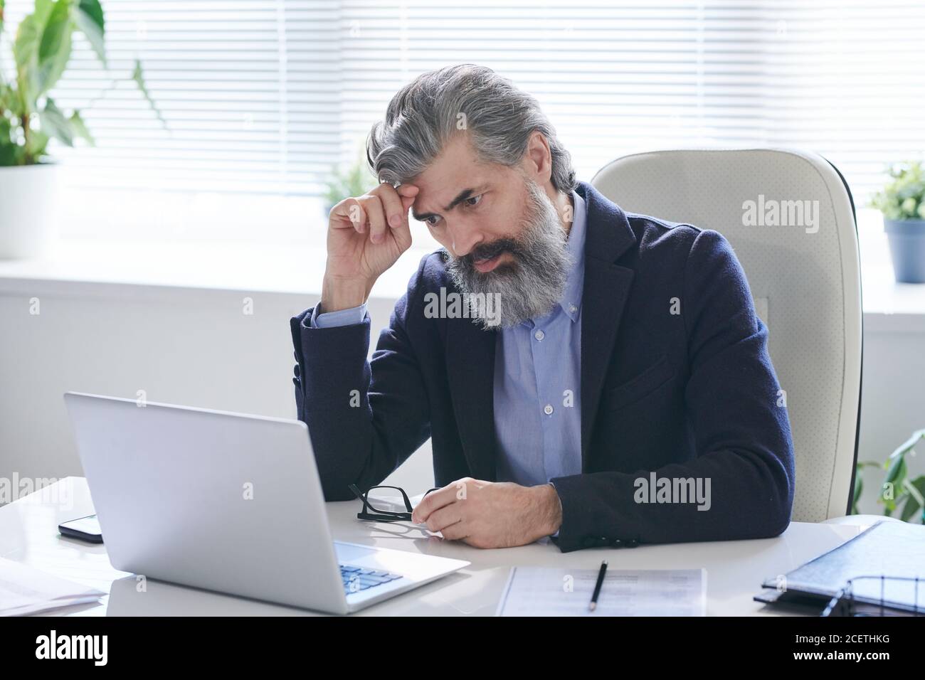 Ritratto di un operaio maschile concentrato che indossa occhiali seduti alla scrivania dell'ufficio e alla lettura delle informazioni sul computer portatile Foto Stock
