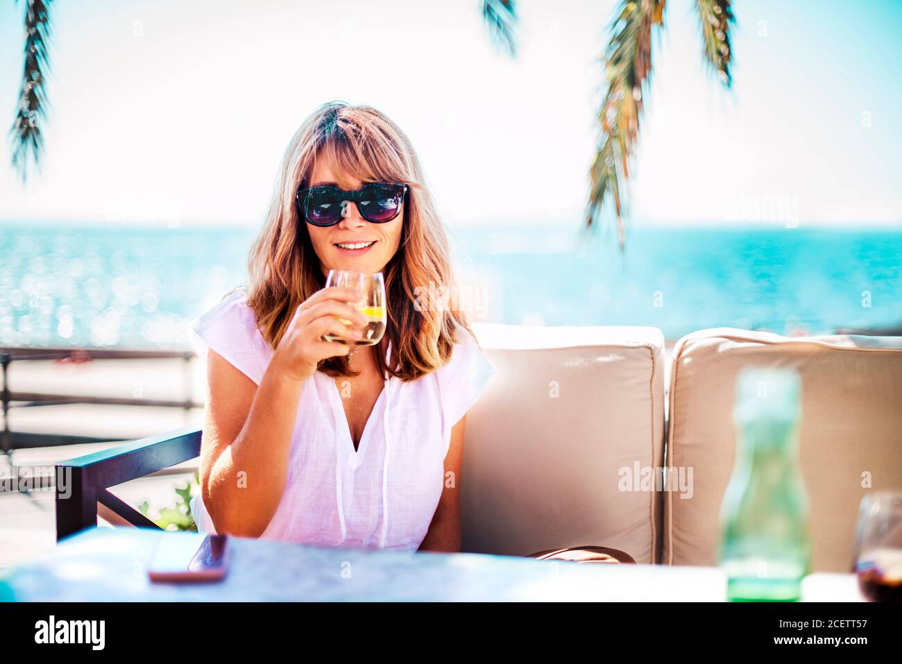 Foto ritratto di donna felice godendo del sole estivo e bevendo un bicchiere d'acqua mentre si rilassa sul divano del salotto vicino al mare. Foto Stock