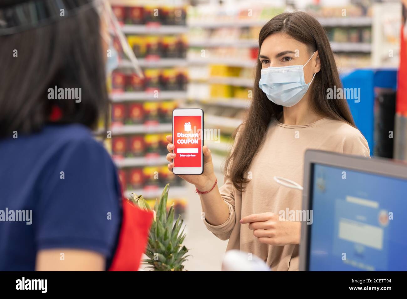 Donna moderna che indossa la maschera sul viso utilizzando un buono sconto digitale sul suo smartphone nel supermercato Foto Stock