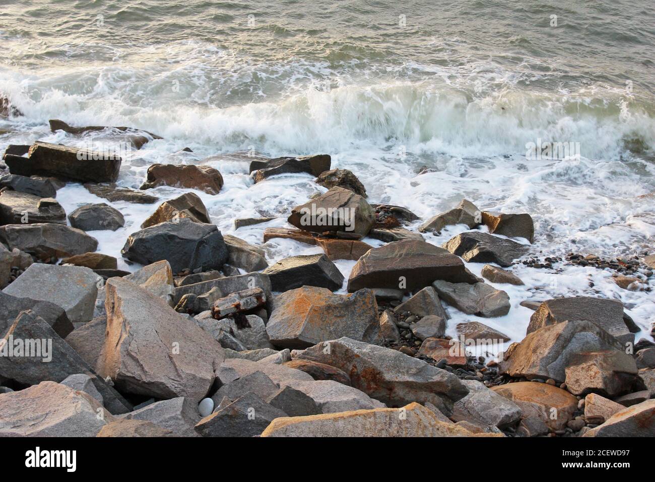 Onde che si infrangono su massi marroni grigi sulla riva di Trefor, Galles del Nord Foto Stock