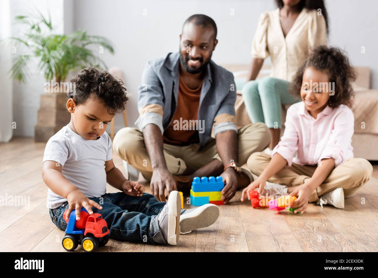 fuoco selettivo dell'uomo afroamericano e dei bambini che giocano con blocchi di costruzione sul pavimento vicino a madre seduta sul letto Foto Stock