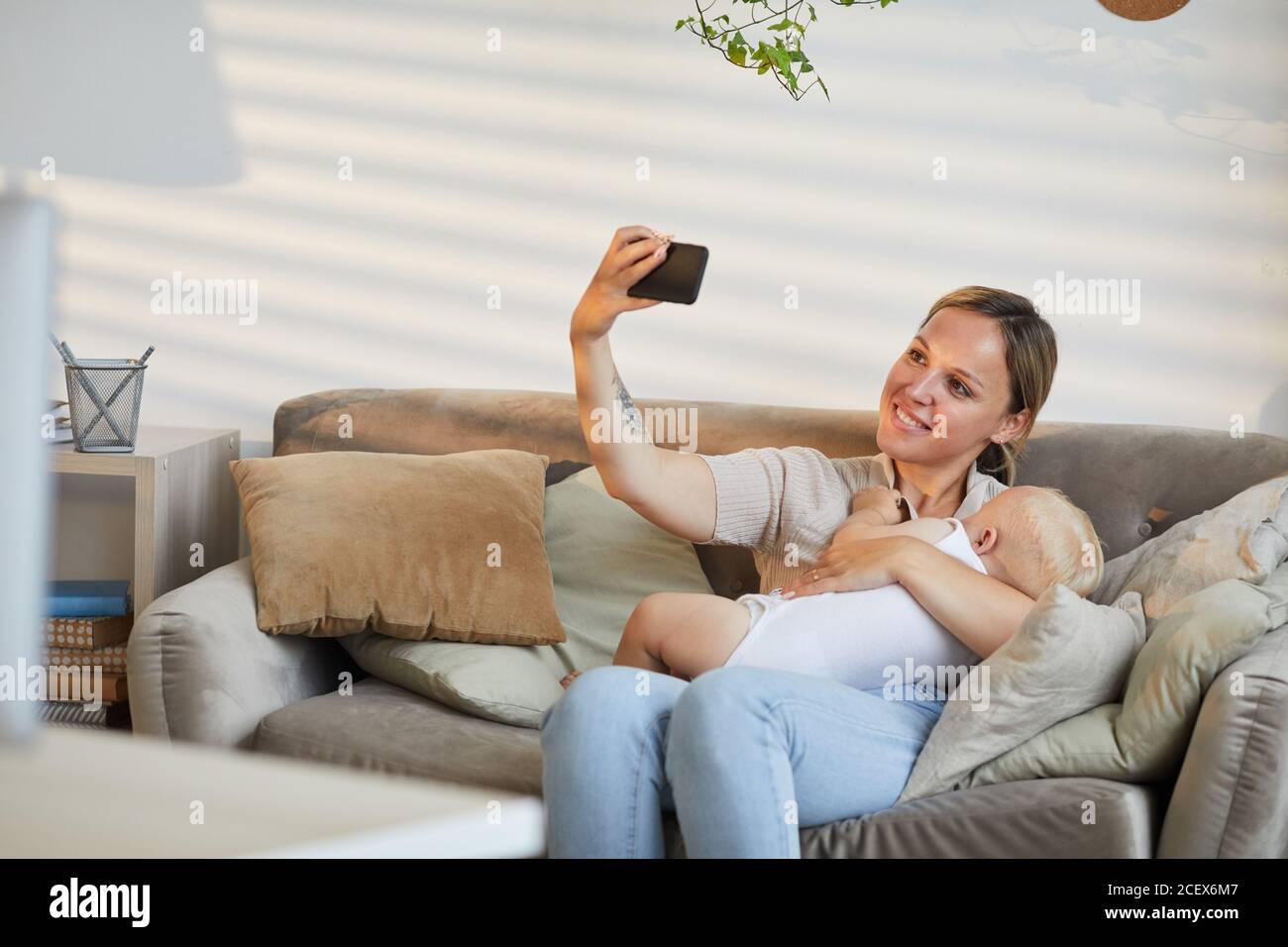 Donna caucasica moderna seduta sul divano scattando foto selfie sopra smartphone mentre coccola il suo bambino tra le braccia Foto Stock