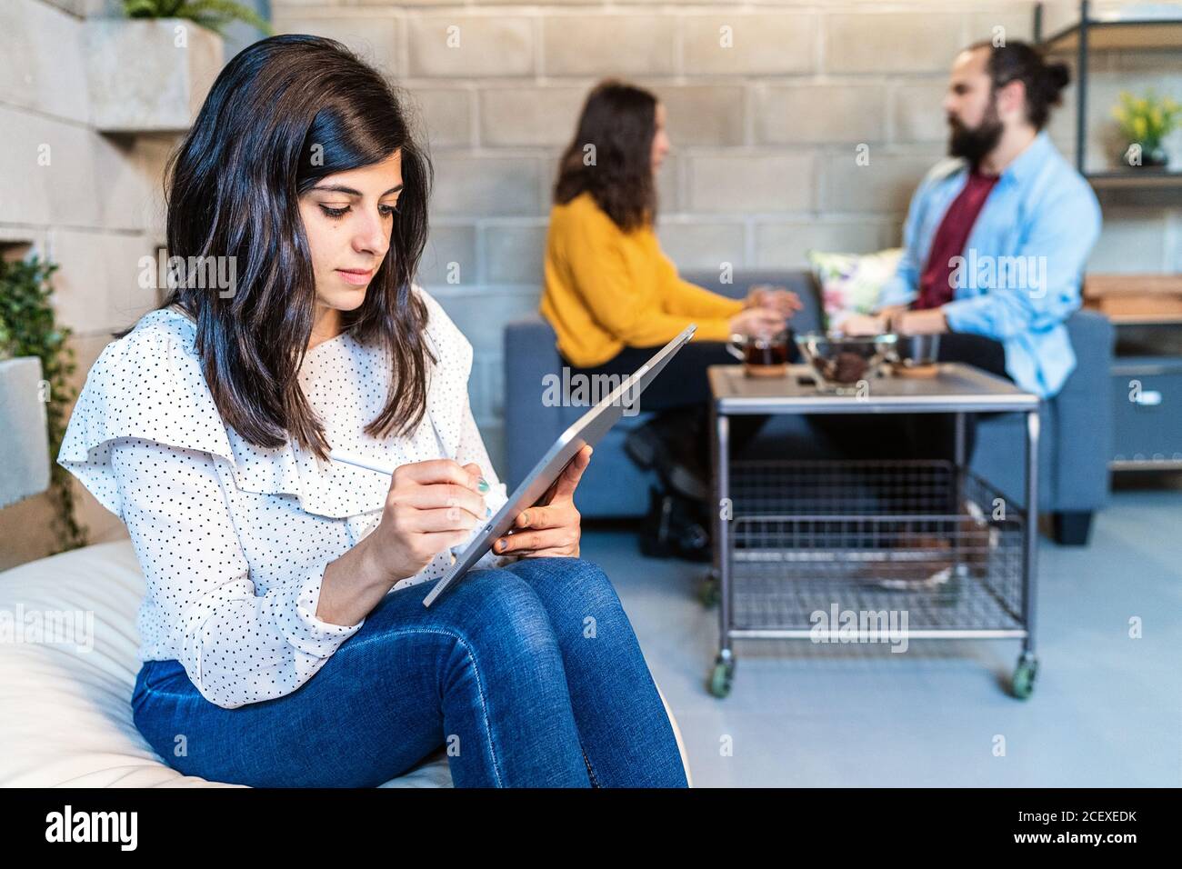 Seria giovane donna designer in abito casual che lavora sul progetto con un moderno tablet grafico mentre sei seduto in un'area di lavoro creativa con colleghi in background Foto Stock