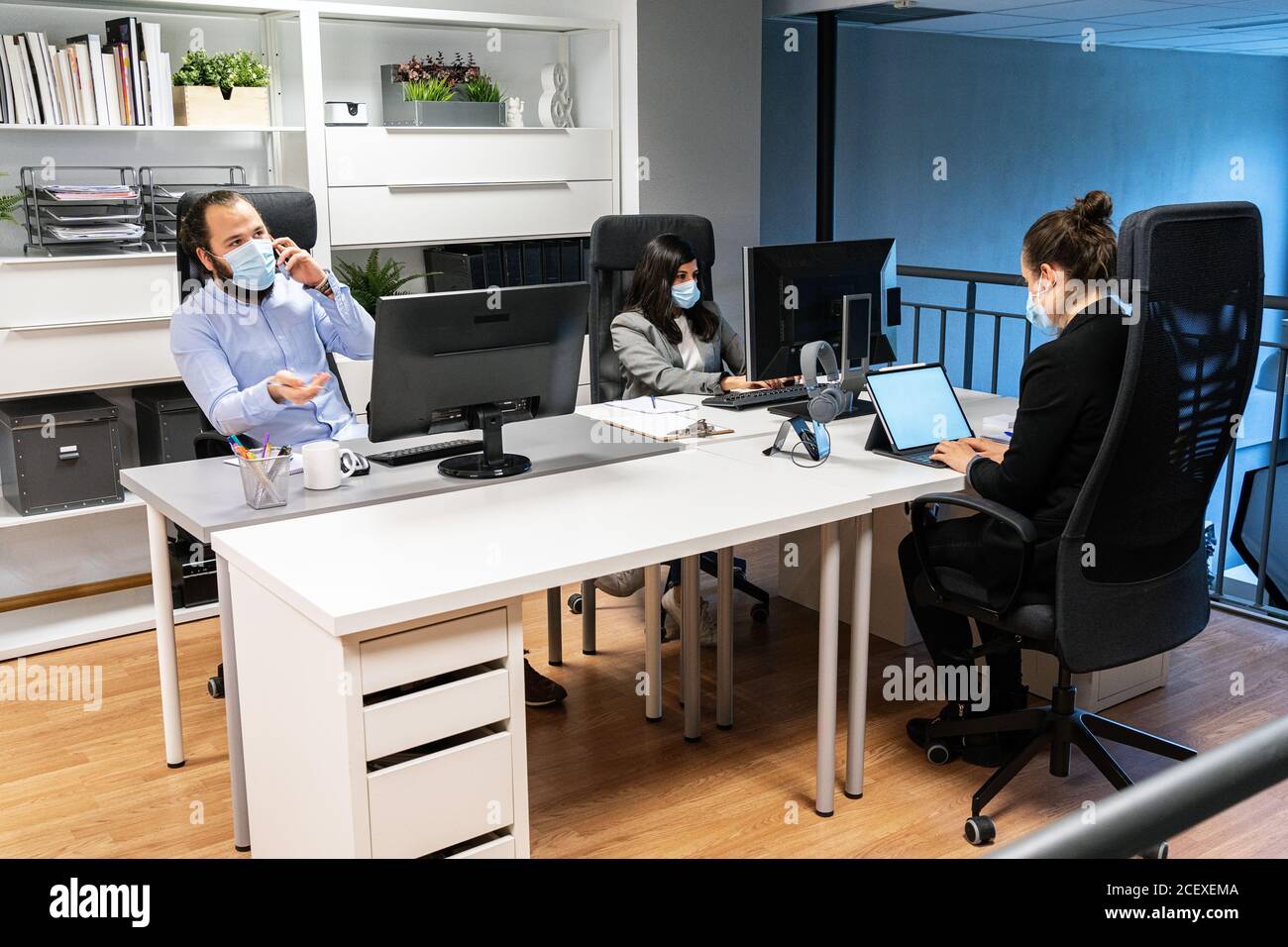 Serio giovane uomo in maschera medica che discute di problema di affari vicino telefono mentre le colleghe femminili lavorano con i calcolatori nel cooworking contemporaneo ufficio Foto Stock