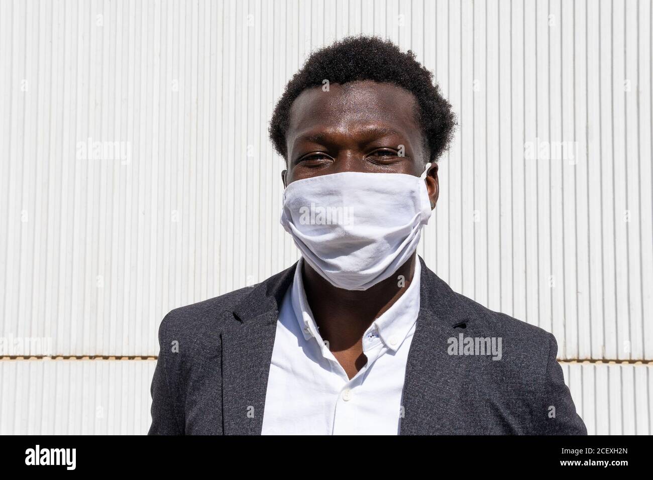 Positivo giovane imprenditore afro-americano in abbigliamento formale e protettivo maschera guardando la macchina fotografica mentre si sta contro il muro sulla strada Foto Stock