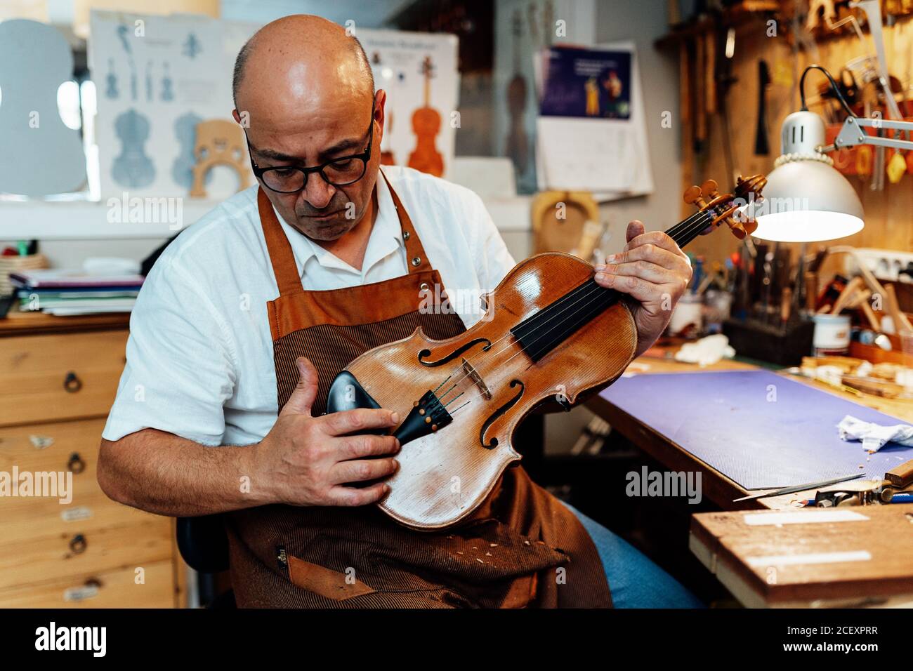 Liutaio maschile maturo in grembiule e bicchieri seduti sulla sedia e tenendo in mano il violino restaurato mentre si lavora in officina Foto Stock