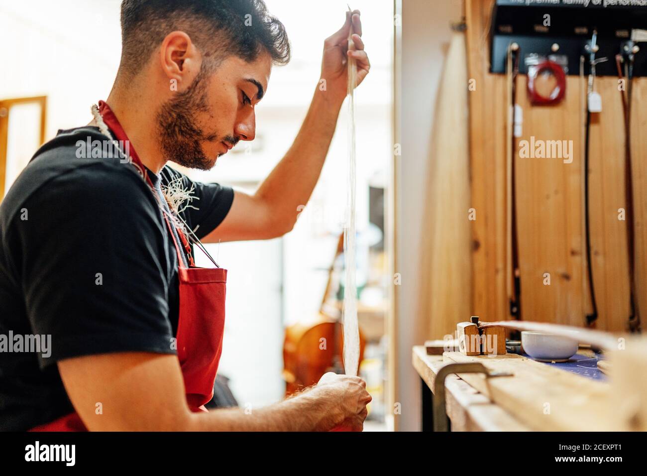 Vista laterale di un giovane maestro artigiano maschile che prepara il fianco di crine per la realizzazione di un arco di violino in officina Foto Stock