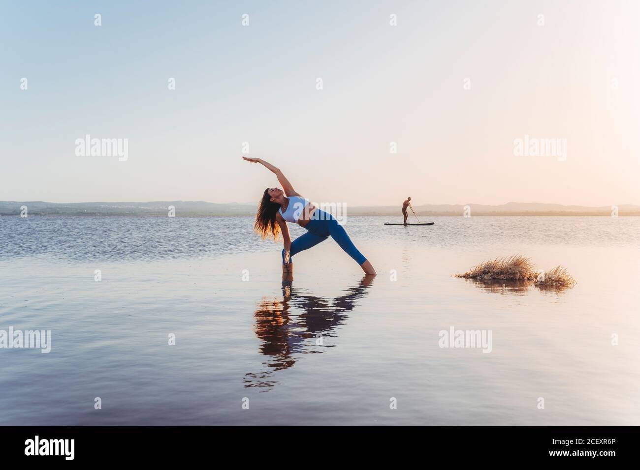 Corpo pieno di femmina sottile irriconoscibile in abbigliamento sportivo che si allunga Extended Side Angle yoga asana durante la pratica sulla riva del lago contro il cielo del tramonto senza nuvole con uomo lontano che naviga in pagaia bordo in background Foto Stock