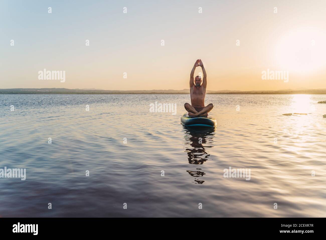 Corpo pieno di uomo messo a fuoco seduto in mezzo posa di loto con le braccia sollevate su tavola galleggiante in un lago calmo acqua contro il cielo del tramonto in estate sera Foto Stock
