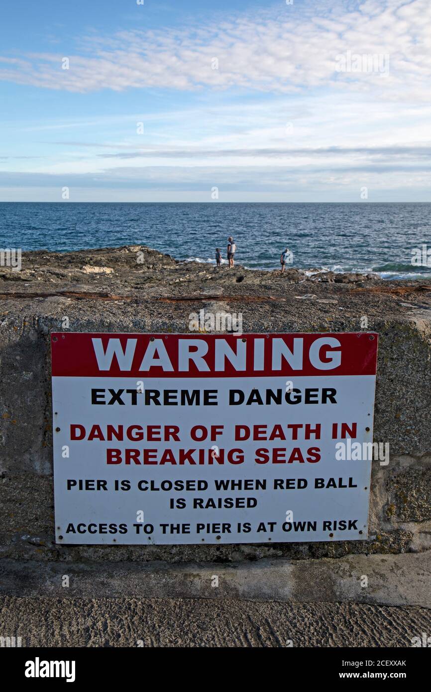 Un segno di avvertimento dei pericoli delle onde e delle maree su una spiaggia in Cornovaglia, Inghilterra, con la gente sulle rocce al bordo delle acque. Foto Stock