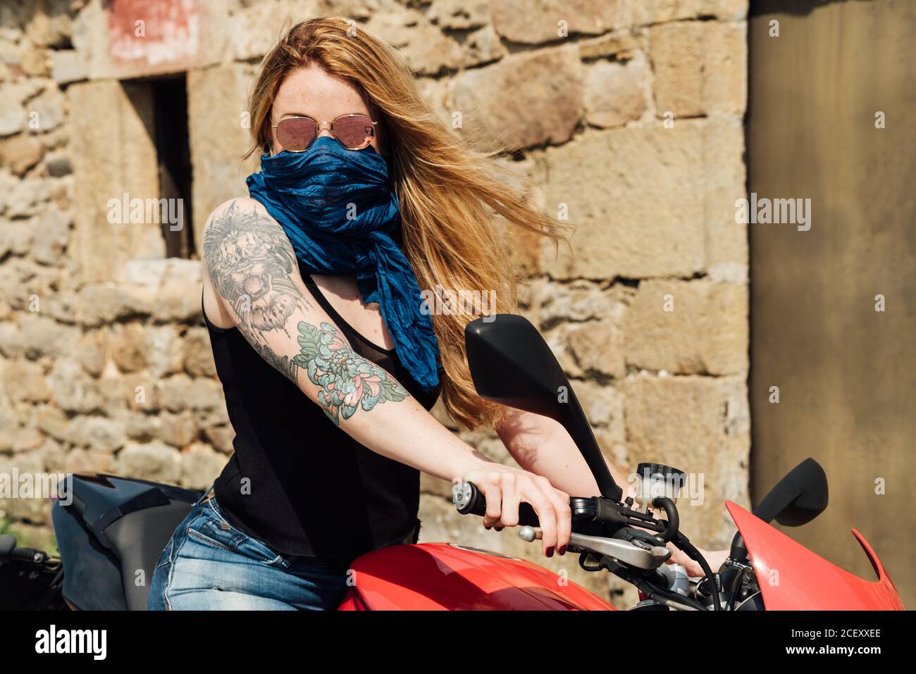 Brutale motociclista femminile con tatuaggi seduti sulla moto moderna mentre copri il viso con la sciarpa e guarda la fotocamera Foto Stock