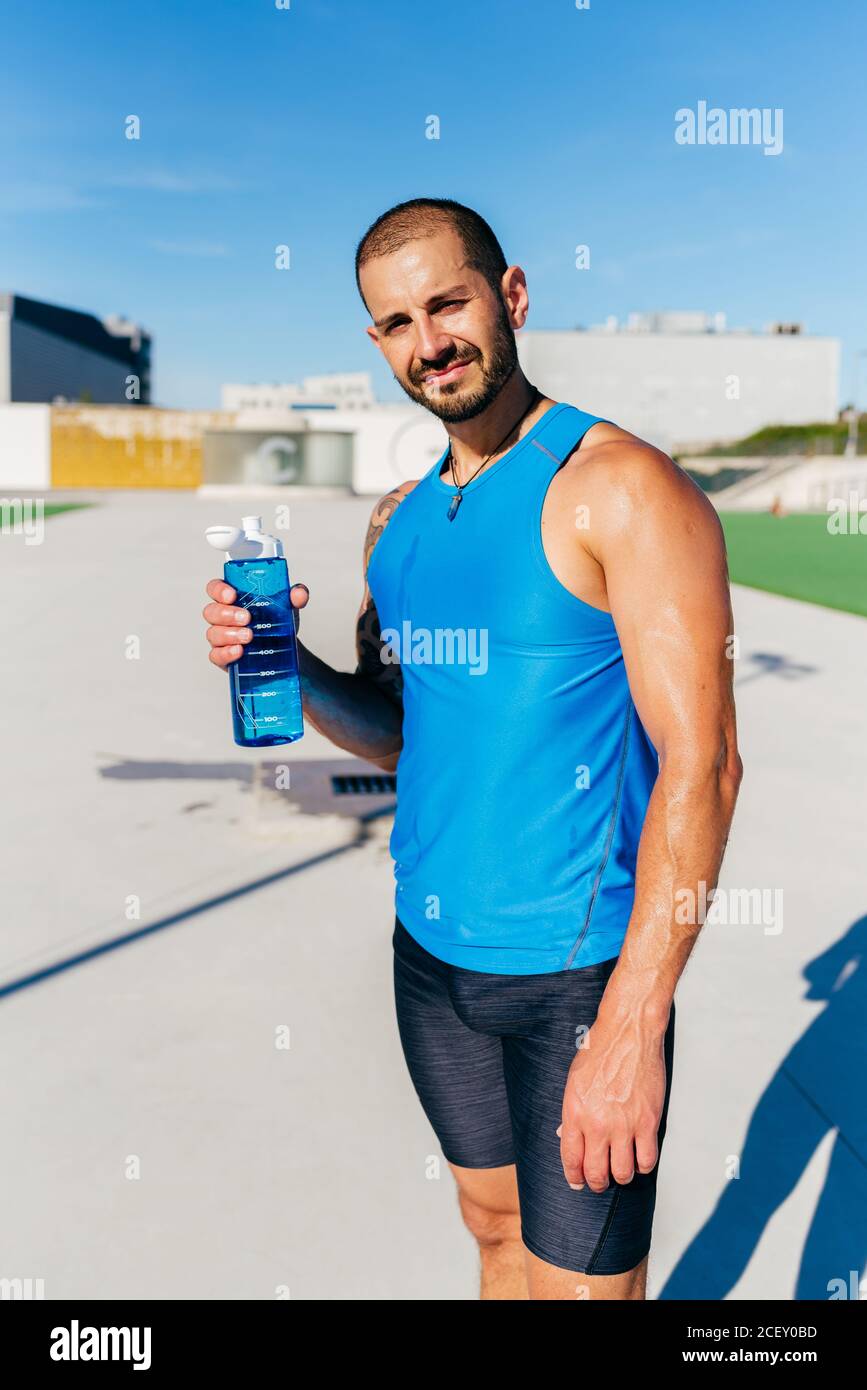 Muscoloso tatuato sudato atleta maschile bere acqua da bottiglia di plastica mentre rinfrescati dopo un intenso allenamento sulla strada urbana Foto Stock