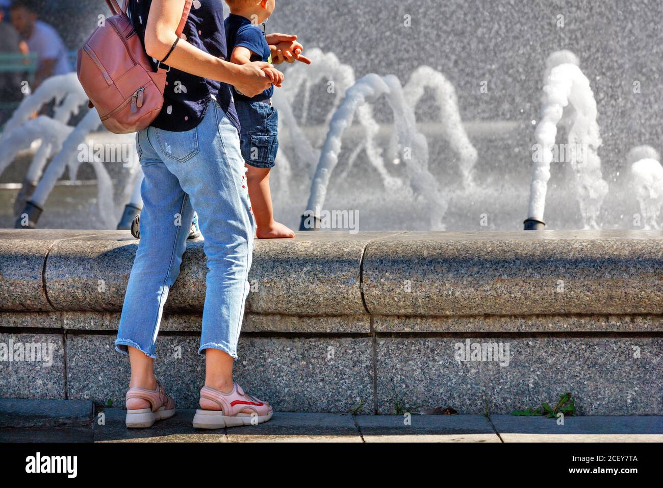 Una madre con un piccolo bambino si trova alla fontana della città nel caldo estivo per rinfrescare. Foto Stock