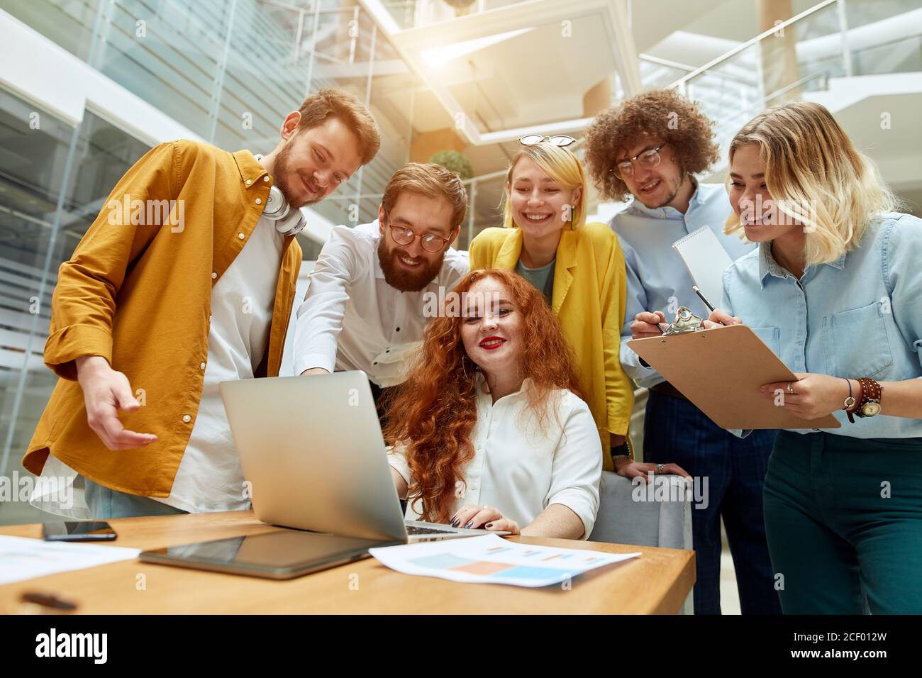 Affascinante donna rossa testa con lunghi capelli ondulati, si siede di fronte al computer portatile, guarda dritto alla fotocamera, sorride ampiamente, mostra faccia felice, positivo a. Foto Stock