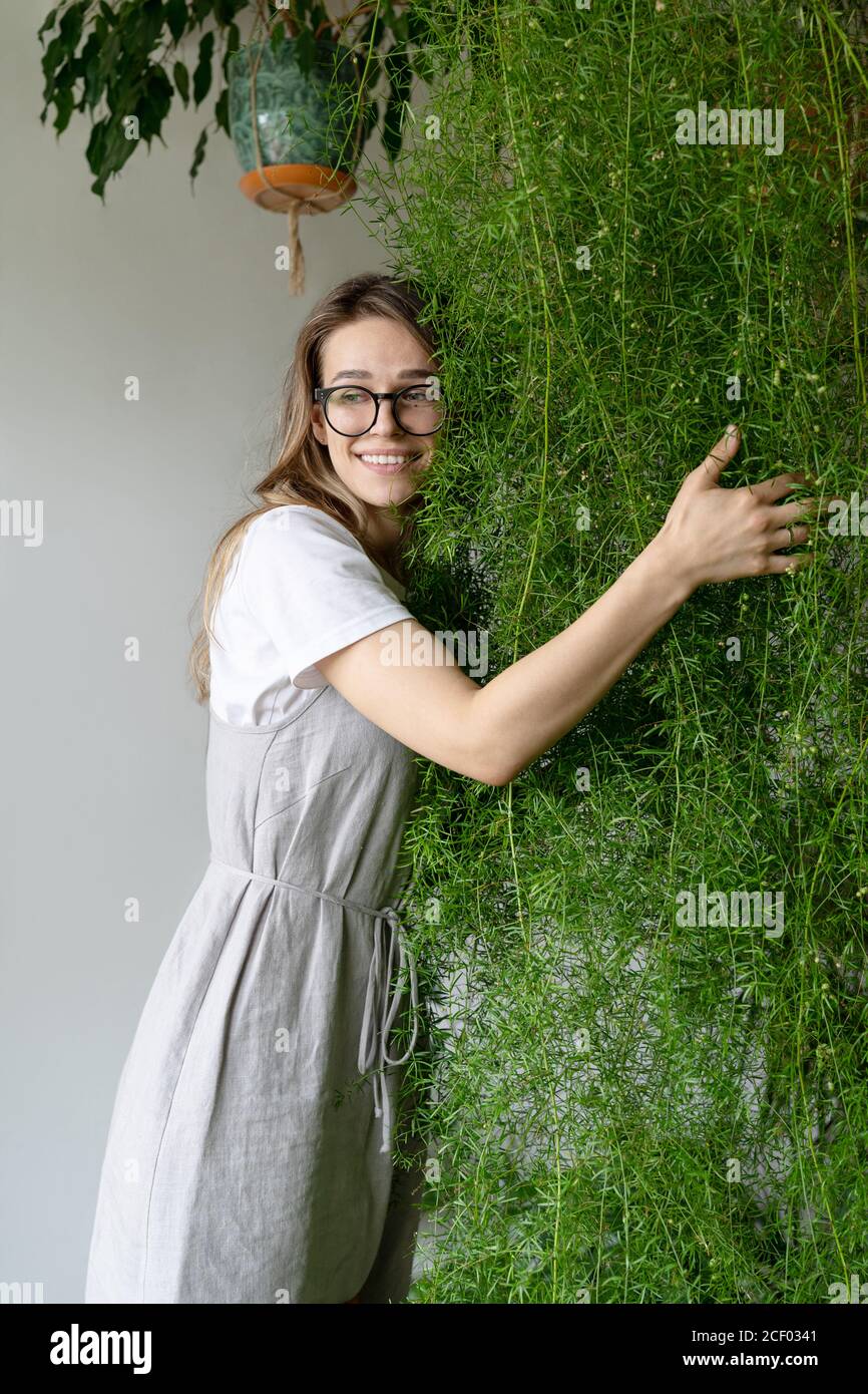 Felice giovane donna giardiniere in abito lieno, abbracciando lussureggiante asparagi felce pianta domestica nel suo negozio di fiori. Verde a casa. Amore delle piante. Interno accogliente Foto Stock