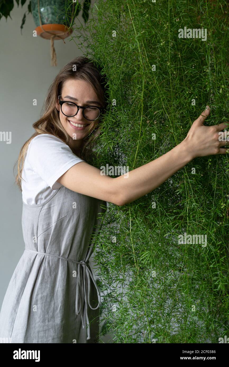 Felice giovane donna giardiniere in abito lieno, abbracciando lussureggiante asparagi felce pianta domestica nel suo negozio di fiori. Verde a casa. Amore delle piante. Interno accogliente Foto Stock