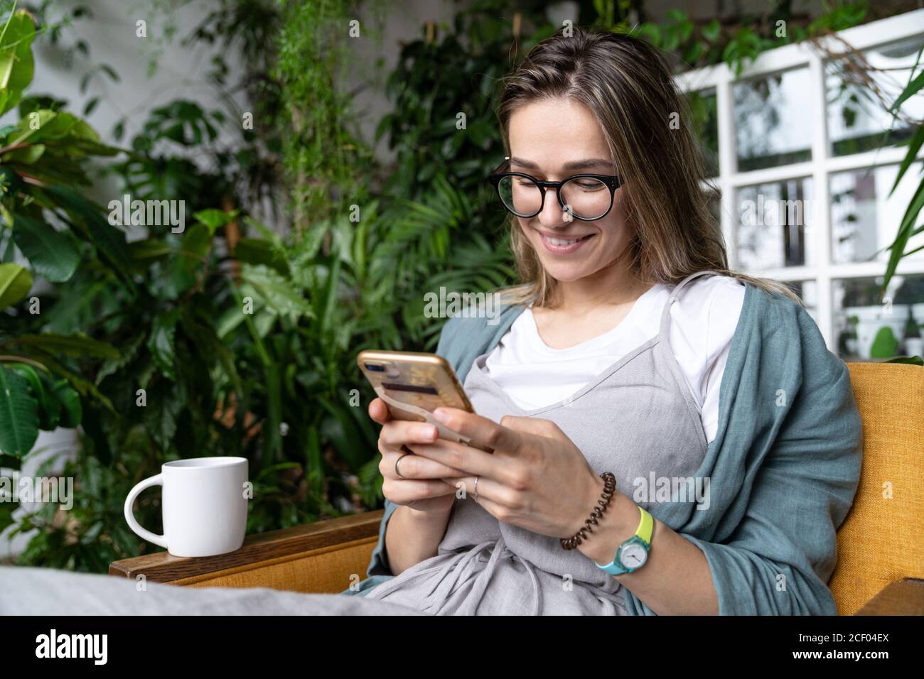 Primo piano di sorridente donna positiva seduta sulla sedia in casa verde, riposo, navigazione internet wireless su smartphone e l'invio di messaggi lei Foto Stock
