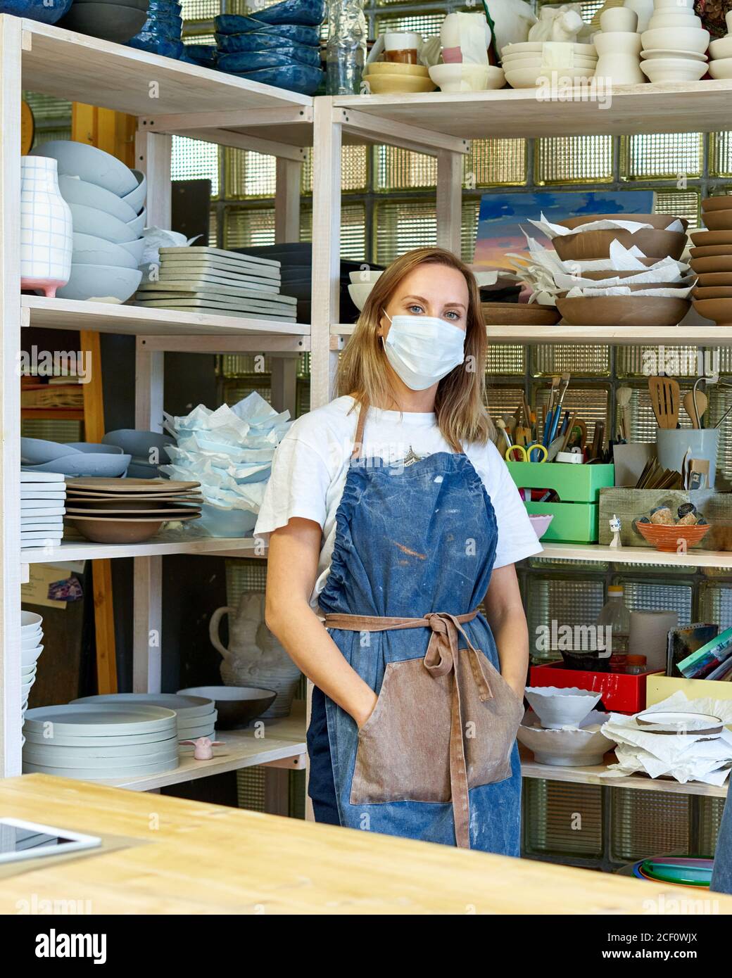 Vita in su ritratto di allegra artigiana femminile con maschera facciale in posa in studio di ceramica in piedi con le braccia incrociate. Nuova normalità con distanza sociale per piccole imprese e imprenditori. Verticale Foto Stock