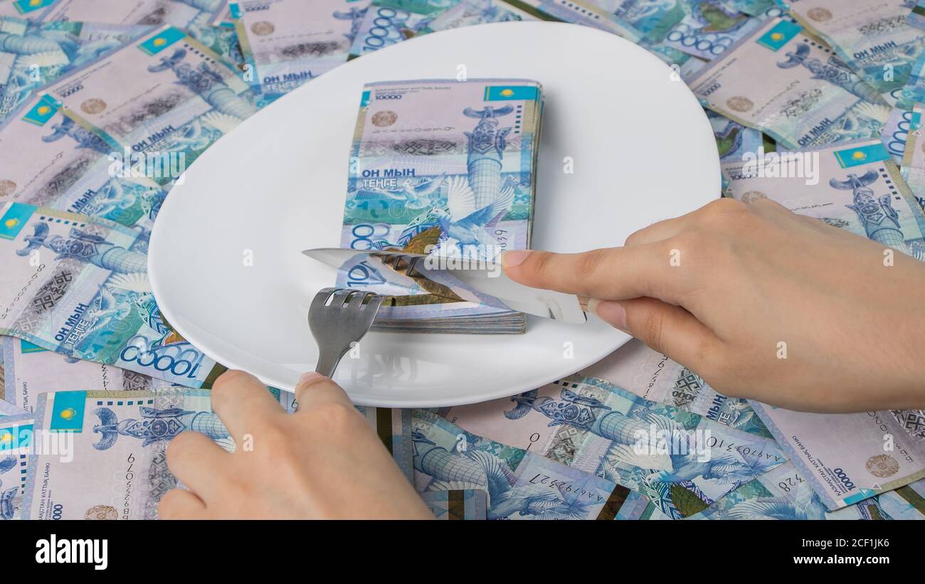 Tagliare il pezzo di denaro in un piatto con un coltello. TENGE, Kazakhstan. Concetto, salario vivente. Economia, Asia centrale. Tagliare il bilancio, dividere, corruttio Foto Stock