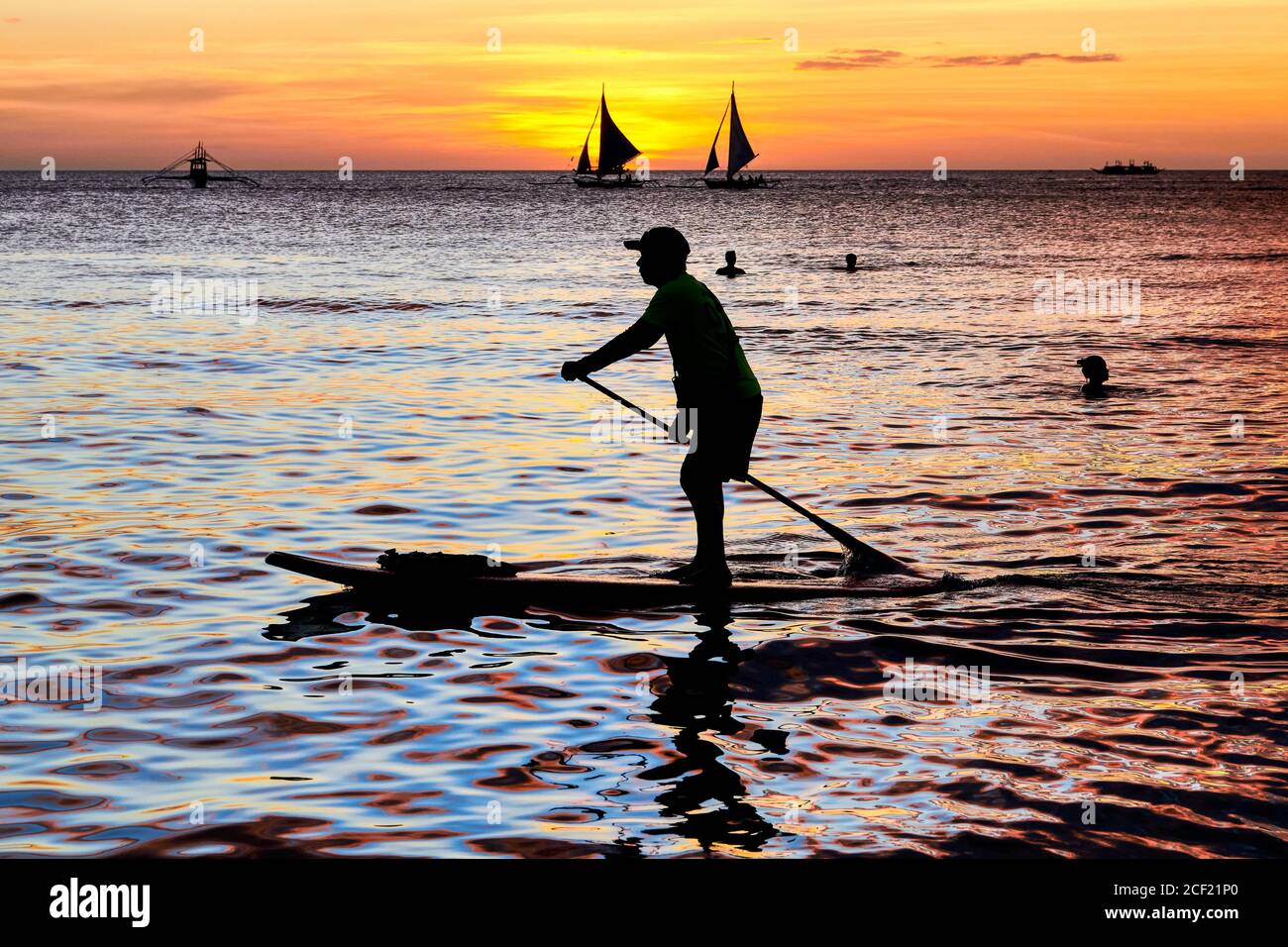 La silhouette di un uomo su una tavola da paddle, la gente che nuota e due barche a vela al tramonto lungo la spiaggia bianca sull'isola di Boracay, Aklan, Filippine. Foto Stock