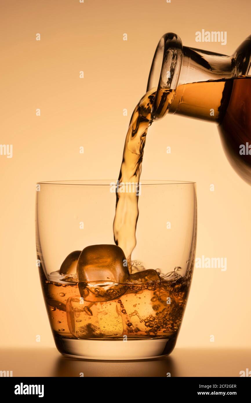 versare whisky o altro alcool in un bicchiere con ghiaccio. primo piano verticale, sfondo arancione Foto Stock