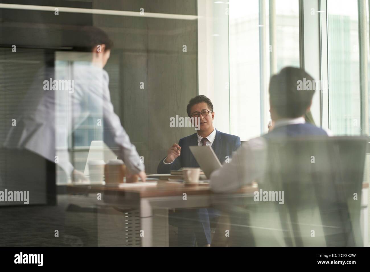 attraverso-la-foto di vetro di gruppo di uomini d'affari asiatici che hanno un discussione in sala riunioni mentre un giovane intern porta il caffè Foto Stock