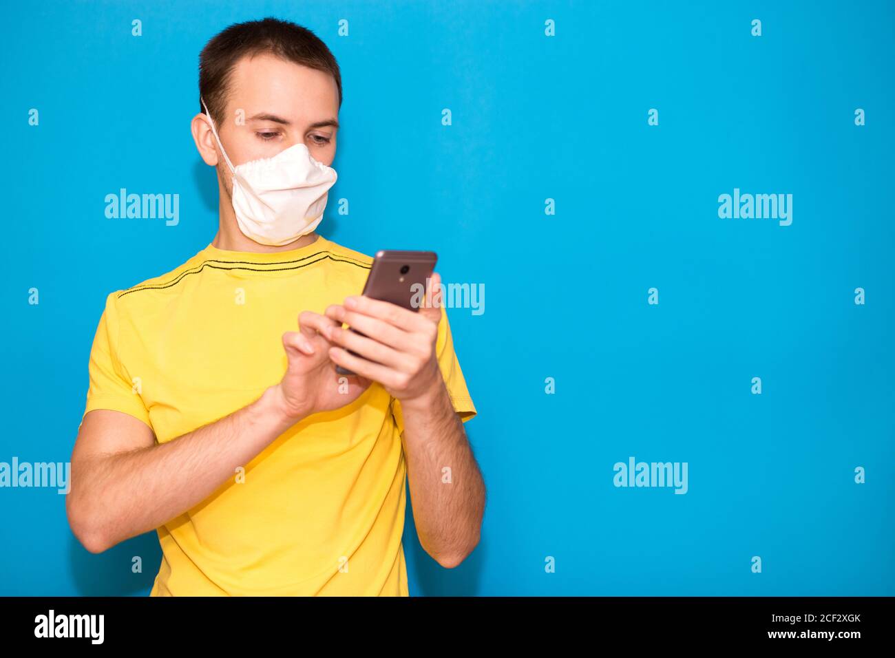 Il giovane indossa maschera medica e t-shirt gialla e l'uso telefono cellulare isolato su sfondo blu Foto Stock