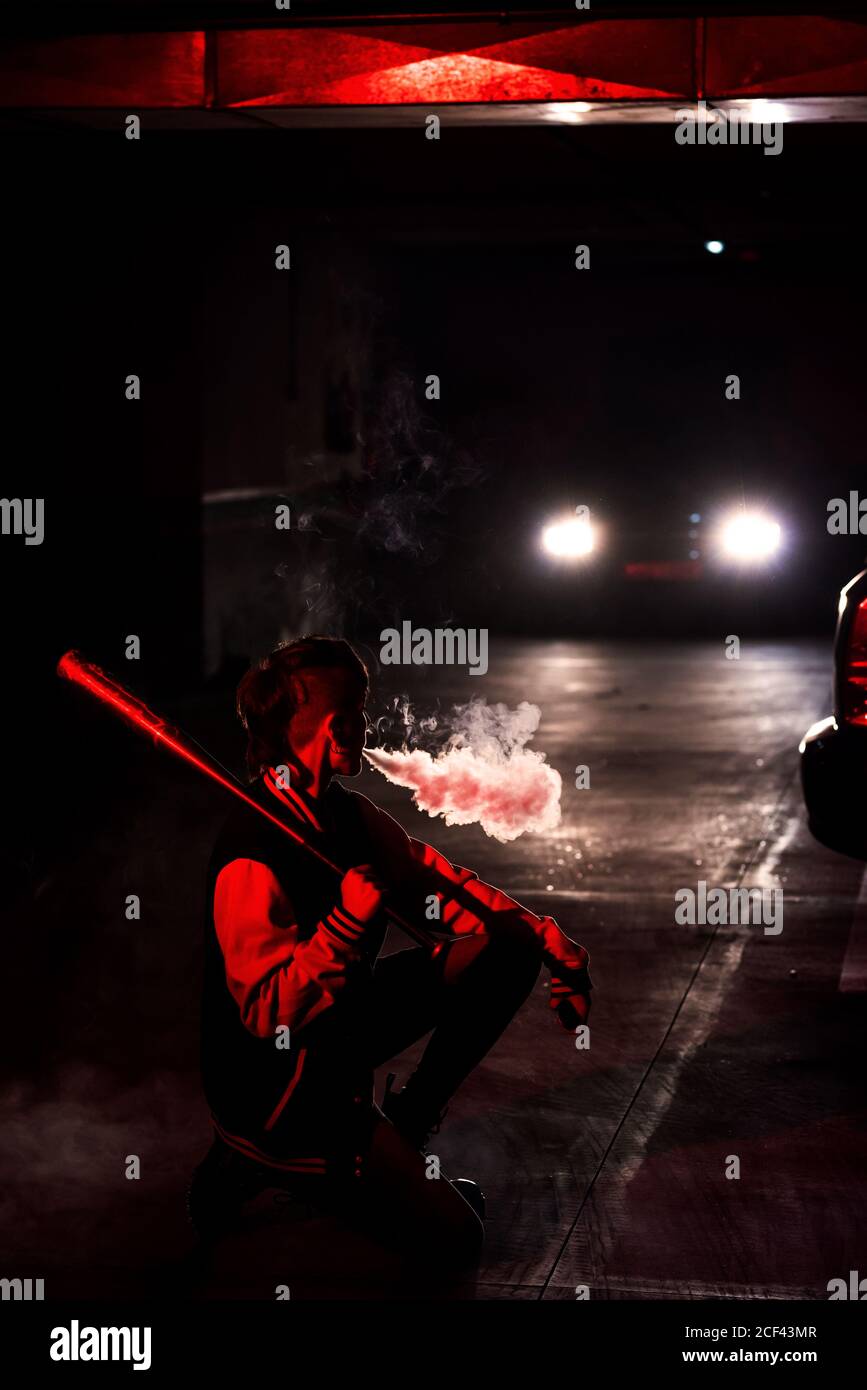 Vista laterale di una donna contemporanea irriconoscibile accovacciata con bomber tenendo una mazza da baseball nera sulla spalla mentre si fuma con luce rossa e luci auto sullo sfondo Foto Stock