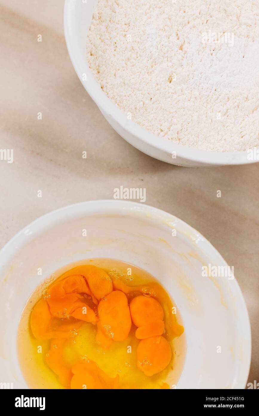 Da sopra ciotole con uova crude e farina di grano messo sul tavolo da cucina durante la preparazione della pasticceria Foto Stock