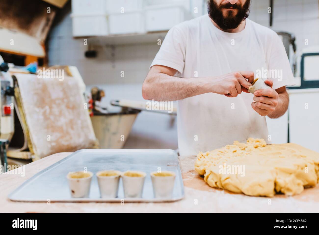 Crop uomo bearded in t-shirt bianca mettendo l'impasto fresco in tazze mentre la preparazione della pasticceria in cucina di panetteria Foto Stock