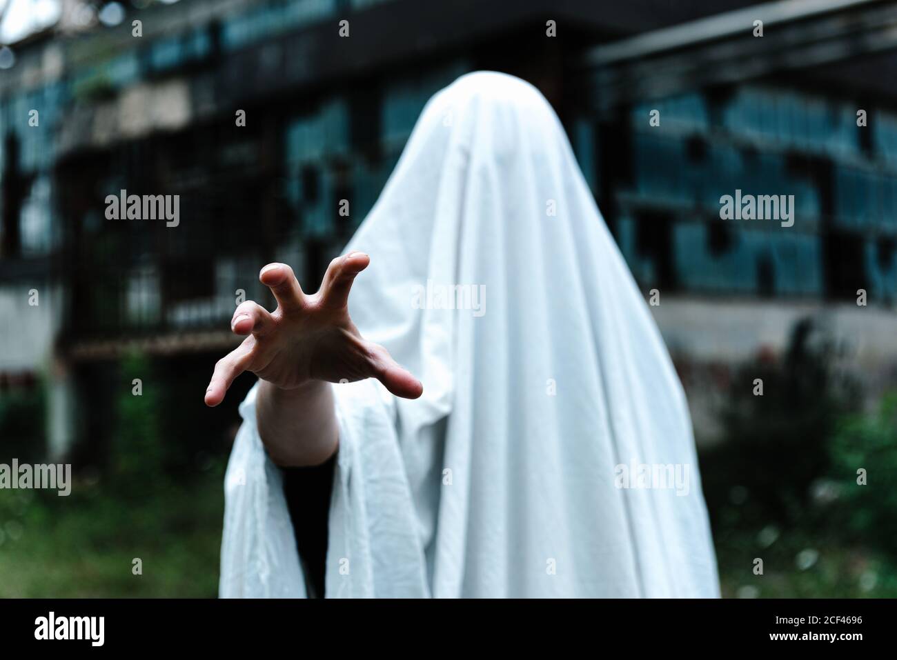 Persona irriconoscibile mano coperta in bianco foglio di letto mascherato come un fantasma vicino ad un vecchio edificio abbandonato con metallo arrugginito costruzione e muri in abda Foto Stock