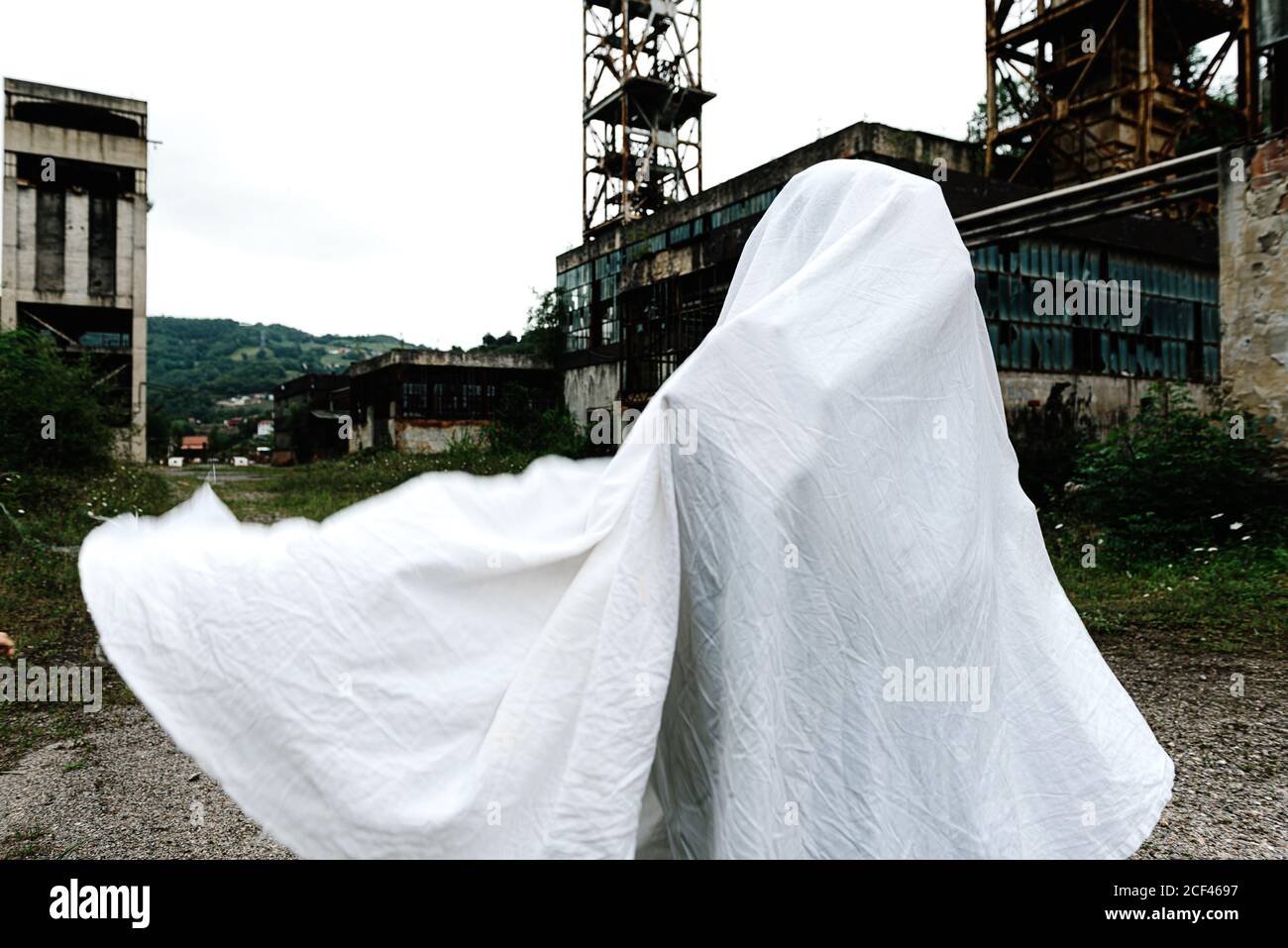 persona irriconoscibile coperta in lenzuolo bianco mascherato come a. ghost vicino vecchio edificio abbandonato con costruzione in metallo arrugginito e muri di scabvicino Foto Stock