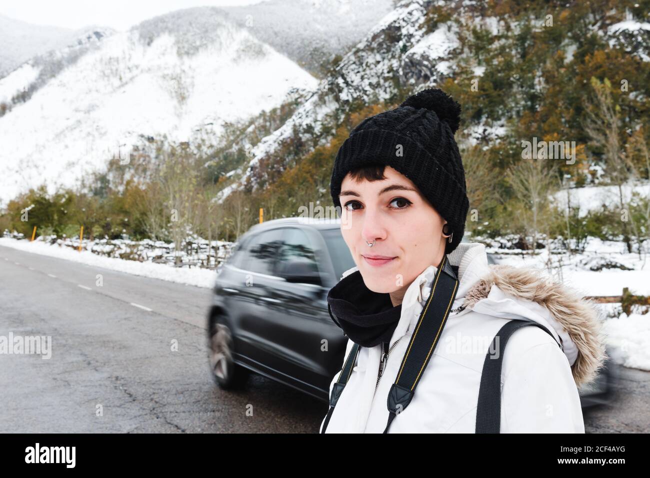 Femmina attiva con piercing al naso in cappello nero e bianco giubbotto invernale in piedi su strada asfaltata con montagne innevate sfondo e guardando la fotocamera Foto Stock