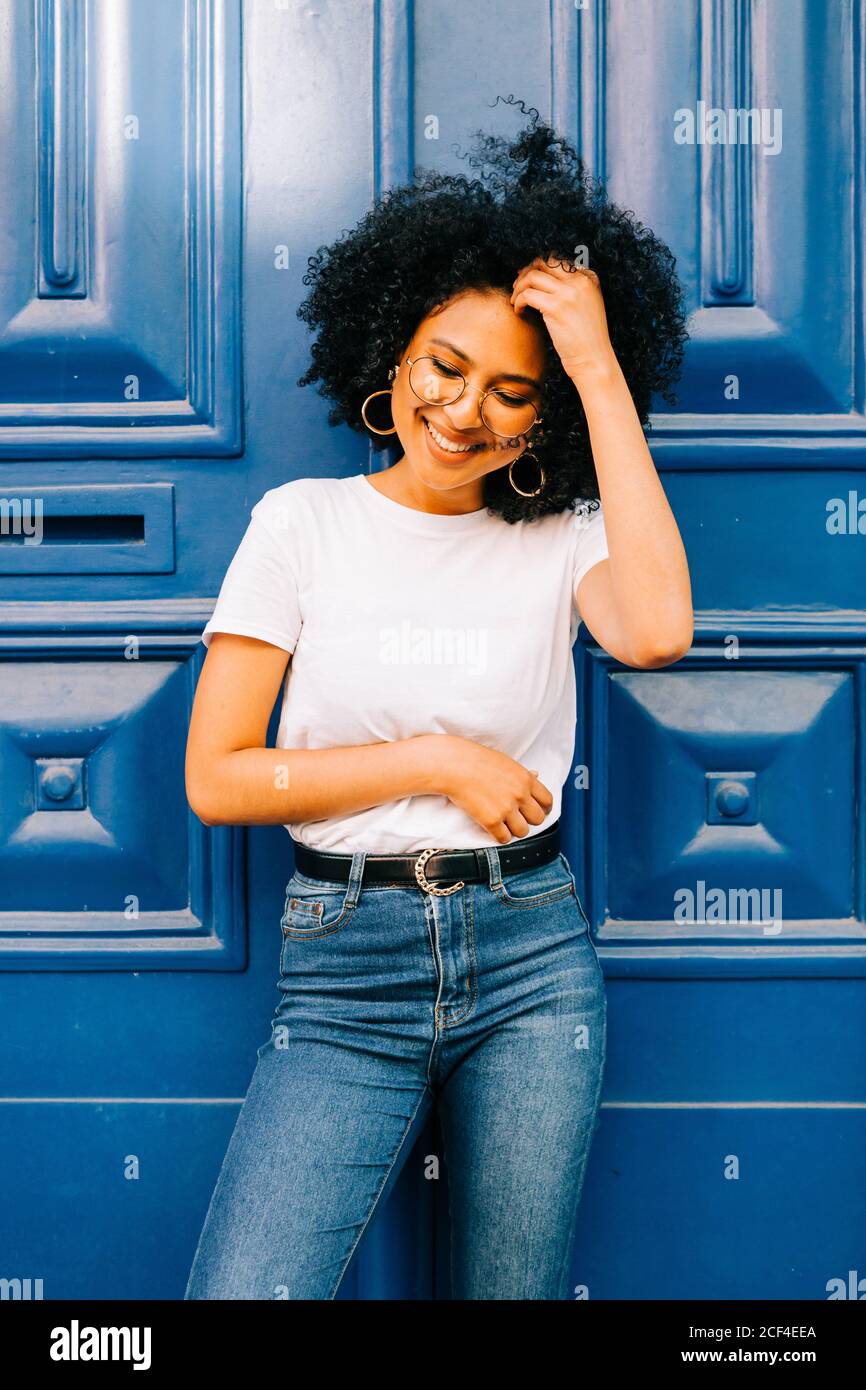 Bella donna etnica in t-shirt bianca e jeans appoggiati su porta blu guardando lontano Foto Stock