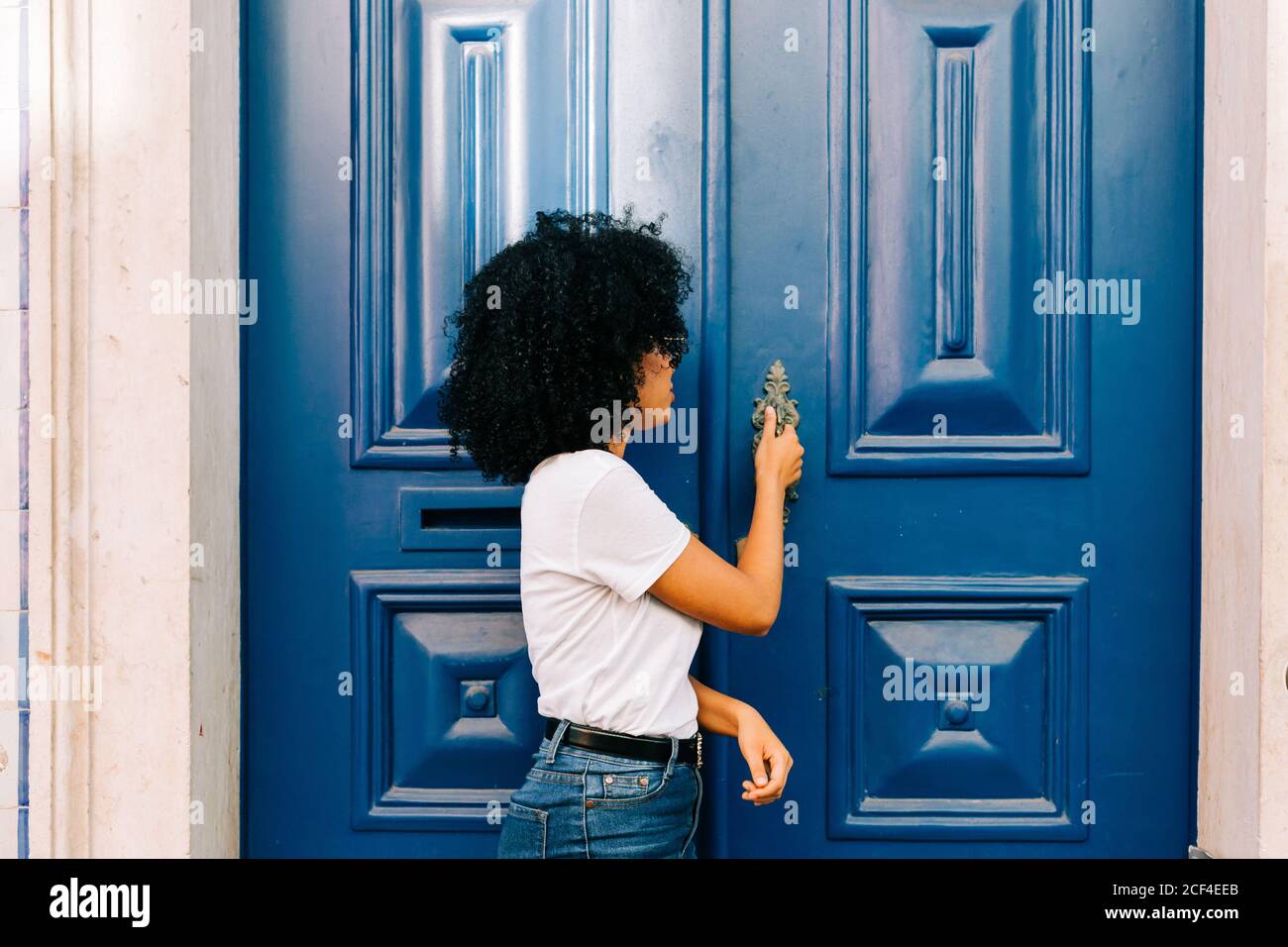 Bella donna etnica in t-shirt bianca e jeans bussando porta blu guardando lontano Foto Stock
