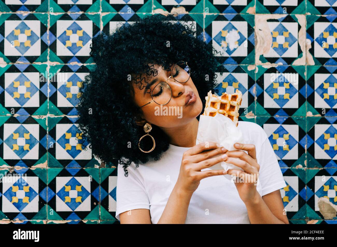 Bella giovane donna in t-shirt bianca in piedi da parete piastrella colorata, mangiare waffle Foto Stock