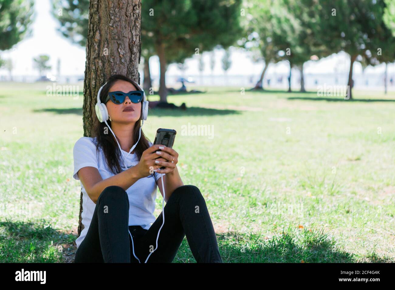 Donna pacifica in occhiali da sole e cuffie con smartphone e ascoltare musica mentre si siede sull'erba nel parco Foto Stock