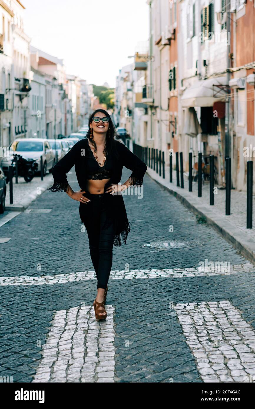 Giovane donna alla moda in vestito nero in piedi sul crosswalk con mani sui fianchi a Lisbona e sorridente Foto Stock