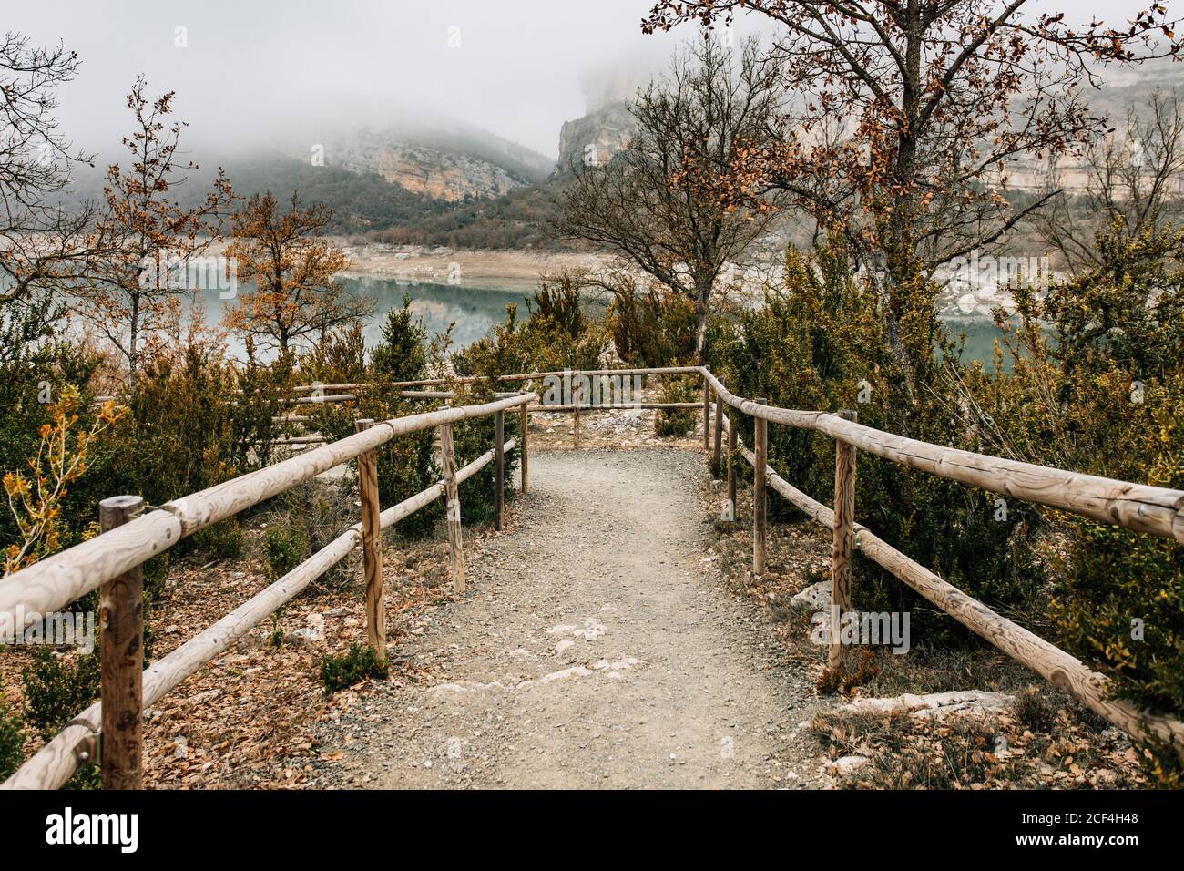 Vuoto percorso curvo con ringhiera di legno che conduce tra gli alberi con fogliame secco sul pendio della collina vicino lago di montagna in nebbia Giorno in Montsec Range in Spagna Foto Stock