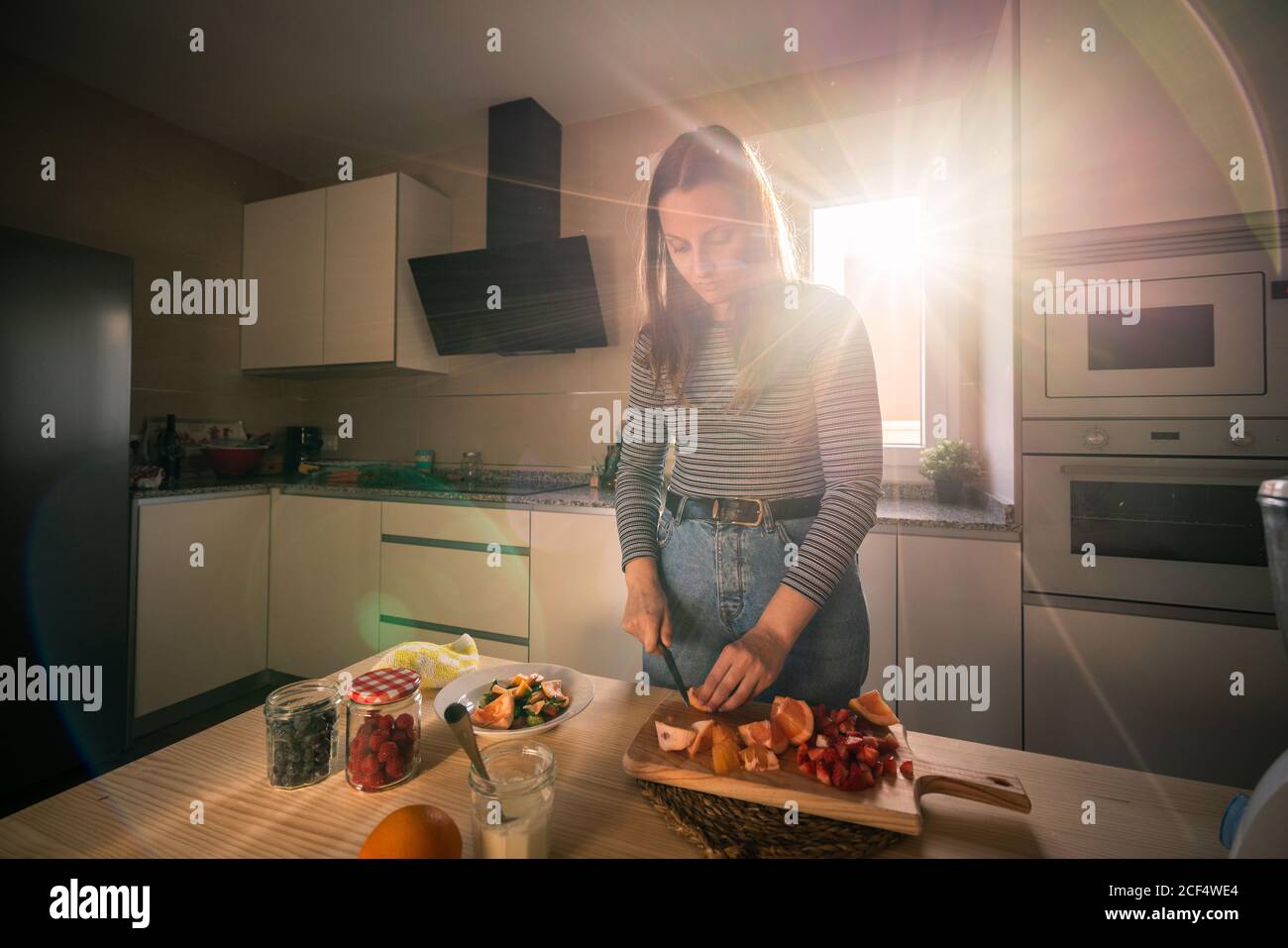 Giovane donna in abito casual tritare frutta fresca durante la cottura in una cucina accogliente sotto travi di luce solare Foto Stock