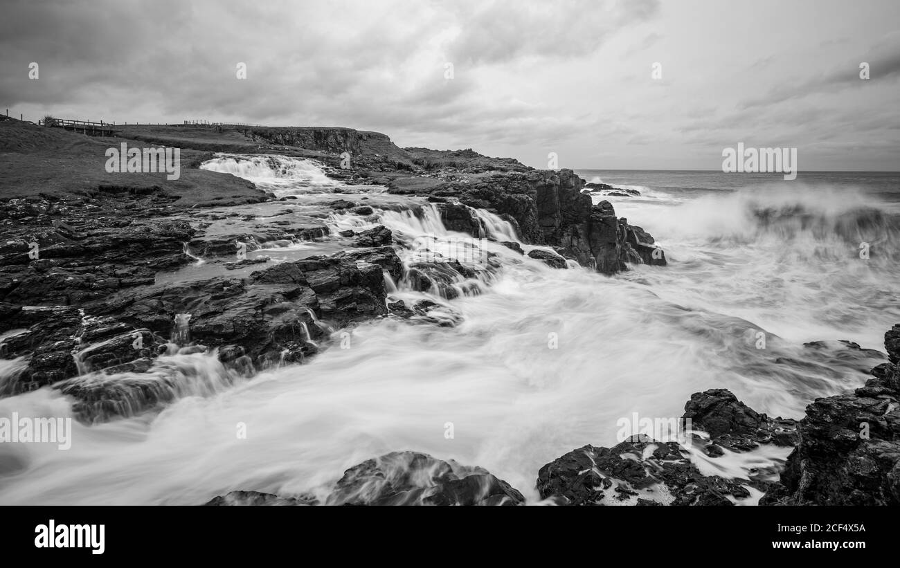 Onde di mare che si infrangono su rocce e si rompono a spruzzi In un giorno tempestoso con nuvole pesanti sulla costa dell'Irlanda del Nord Foto Stock