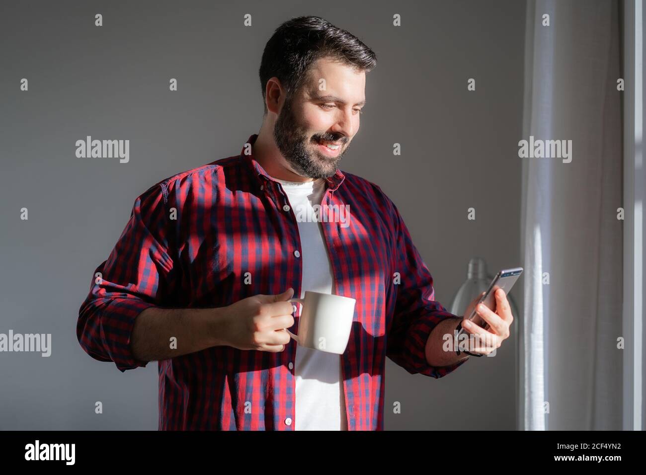 Un giovane sta guardando il telefono cellulare. L'uomo sta lavorando da casa. L'imprenditore sta tenendo un telefono e bevendo da una tazza. Sta sorridendo Foto Stock