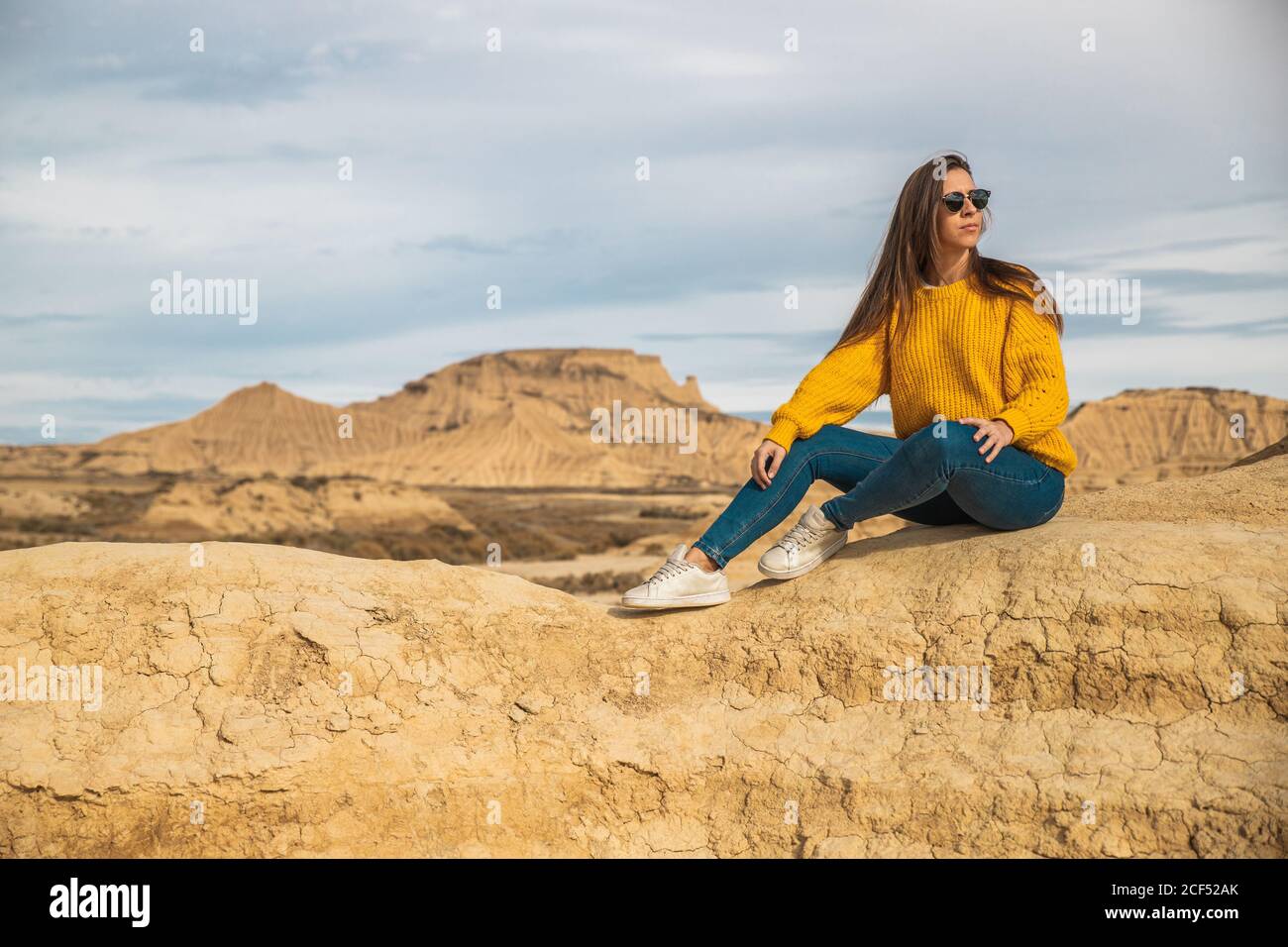 Gioiosa giovane viaggiatore femminile in elegante abbigliamento casual seduto in collina marrone con cielo blu su sfondo a Bardenas Reales, Navarre, Spagna Foto Stock