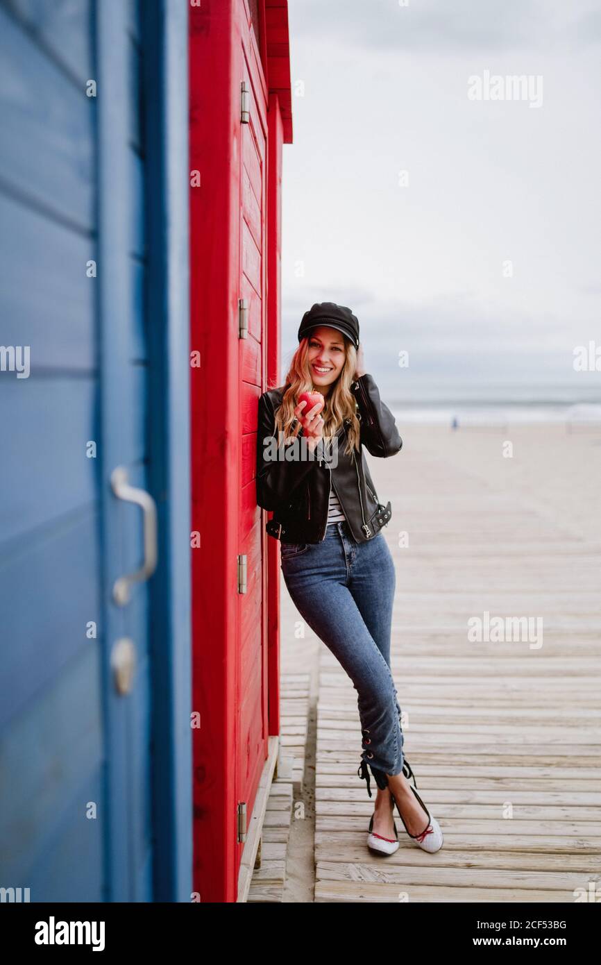 Bionda trendy dai capelli lunghi Donna in berretto nero e giacca di pelle sorridente brillante alla macchina fotografica che mangia una mela rossa mentre si appoggia su una spiaggia di legno cabine Foto Stock