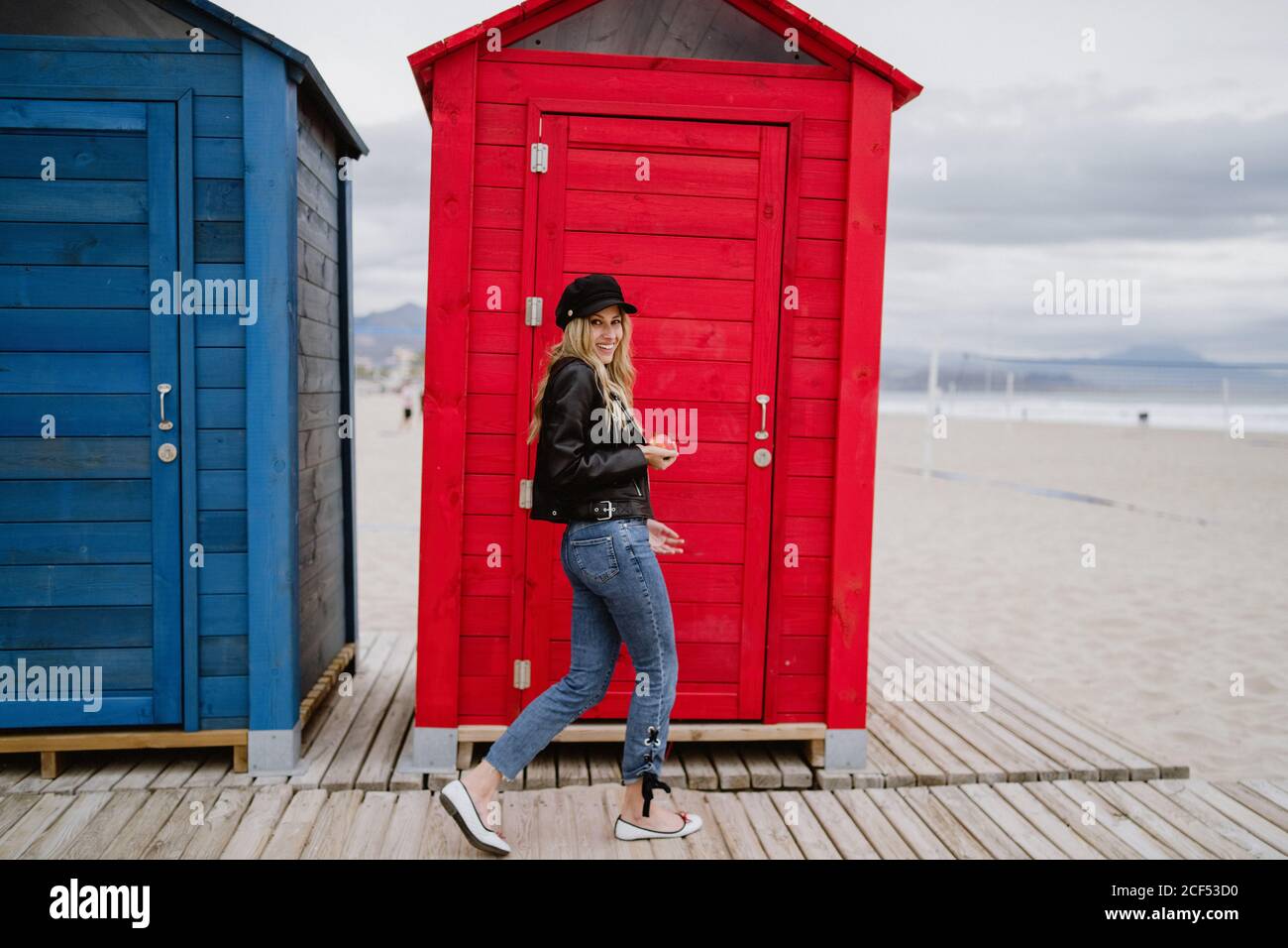 Bionda trendy dai capelli lunghi Donna in berretto nero e giacca di pelle sorridente brillantemente alla macchina fotografica che mangia una mela rossa mentre cammina davanti a una cabina di spiaggia di legno Foto Stock