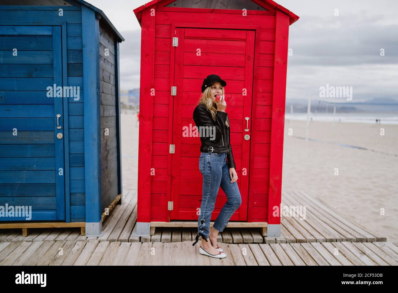 Bionda trendy dai capelli lunghi Donna in berretto nero e giacca di pelle sorridente brillantemente alla macchina fotografica che mangia una mela rossa mentre cammina davanti a una cabina di spiaggia di legno Foto Stock