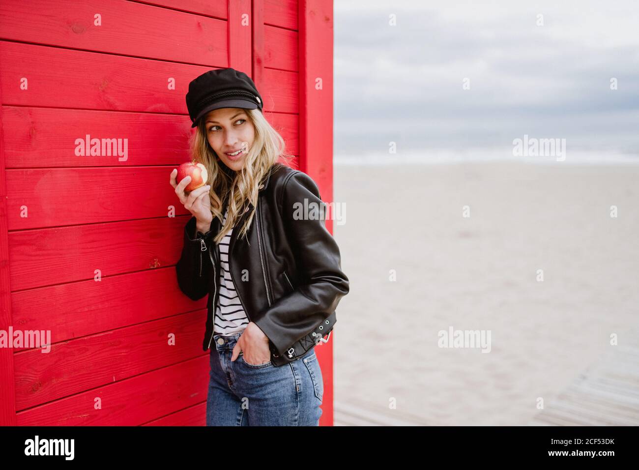 Bionda trendy dai capelli lunghi Donna in berretto nero e giacca di pelle sorridente brillante alla macchina fotografica che mangia una mela rossa mentre si appoggia su una spiaggia di legno cabine Foto Stock